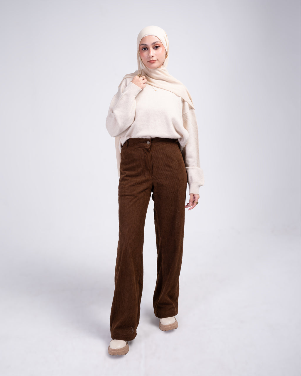 Woman wearing a cream hijab, white top, and brown pants on a white background